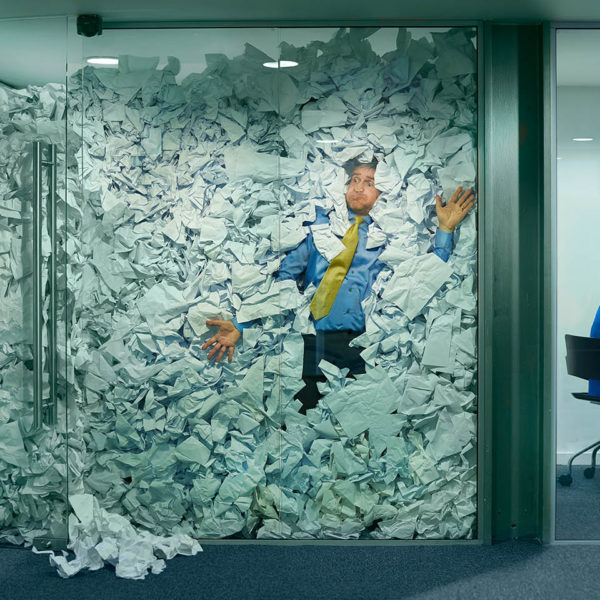 A man squished against a glass wall by a mountain of crumpled paper